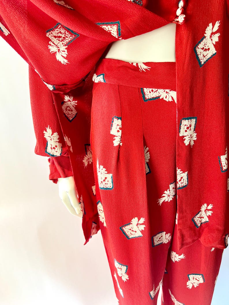May include: A red two-piece outfit featuring a jacket and pants. The fabric is patterned with white floral and geometric designs with teal accents. The jacket has long sleeves and buttons. The pants have a high waist and a straight leg.