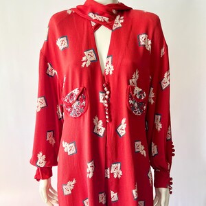 May include: Red vintage jumpsuit with a unique print of white and blue floral patterns. Features a collared neckline, long sleeves, and decorative buttons down the front. The jumpsuit has two front pockets and decorative buttons on the cuffs.