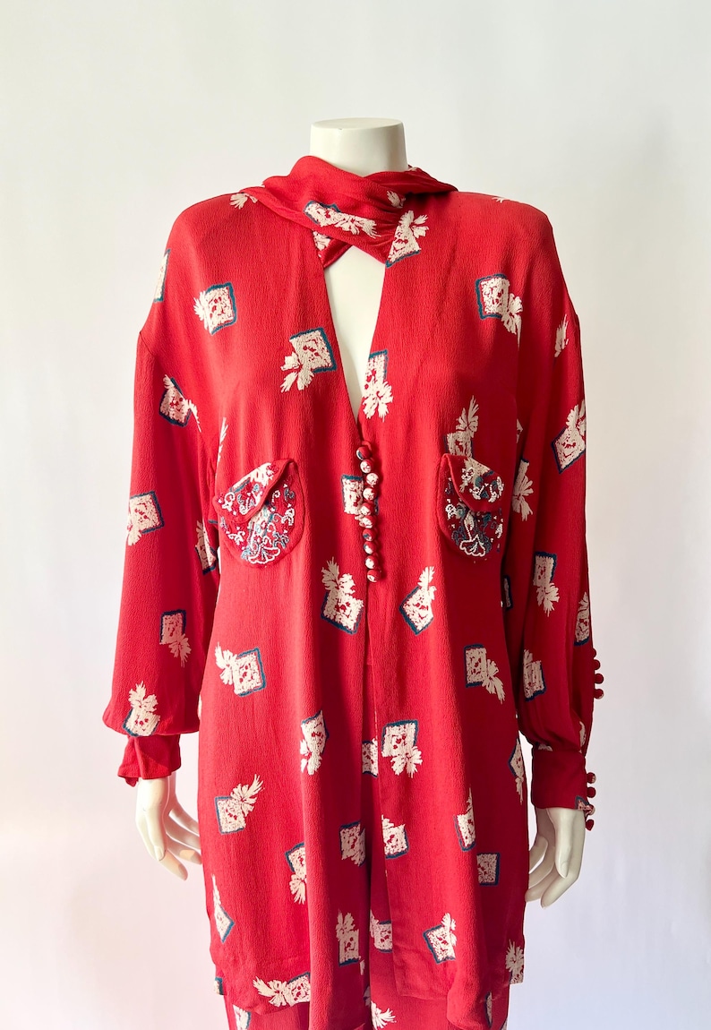 May include: Red vintage jumpsuit with a unique print of white and blue floral patterns. Features a collared neckline, long sleeves, and decorative buttons down the front. The jumpsuit has two front pockets and decorative buttons on the cuffs.