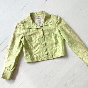 Puede incluir: Chaqueta verde claro con botones y mangas largas. La chaqueta tiene cuello y un sutil estampado de cachemira. La tela tiene un ligero brillo. La chaqueta est&aacute; sobre una superficie de madera blanca.
