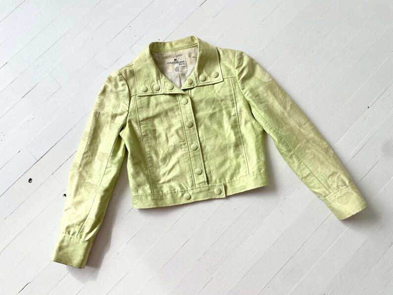 Puede incluir: Chaqueta verde claro con botones y mangas largas. La chaqueta tiene cuello y un sutil estampado de cachemira. La tela tiene un ligero brillo. La chaqueta est&aacute; sobre una superficie de madera blanca.