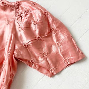 May include: A pink satin blouse with a floral embroidered pattern. The blouse has short, puffed sleeves and a gathered neckline.