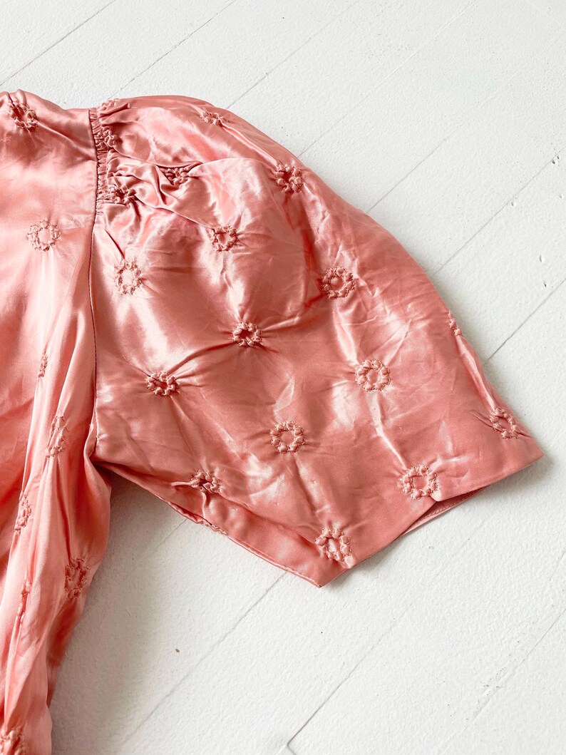 May include: A pink satin blouse with a floral embroidered pattern. The blouse has short, puffed sleeves and a gathered neckline.