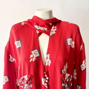 May include: A red vintage blouse with a unique collar and button closure. The blouse features a repeating pattern of white floral and square designs with teal accents. Two small pockets with embroidered details are visible.
