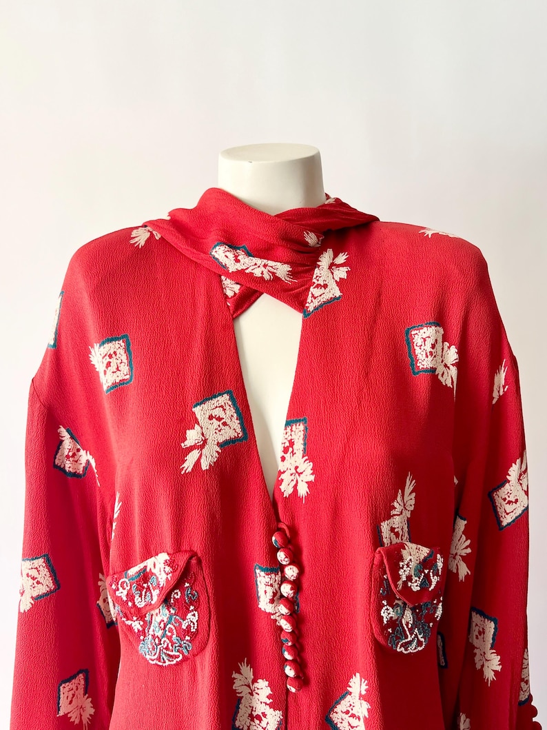 May include: A red vintage blouse with a unique collar and button closure. The blouse features a repeating pattern of white floral and square designs with teal accents. Two small pockets with embroidered details are visible.