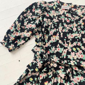 以下が含まれることがあります： ピンク、白、緑の花柄の黒い花柄ドレス。 ドレスはウエストにリボン、プリーツのボディス、パフスリーブが特徴です。 ドレスはヴィンテージ感があります。