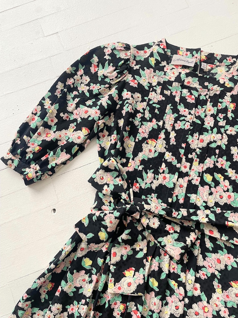以下が含まれることがあります： ピンク、白、緑の花柄の黒い花柄ドレス。 ドレスはウエストにリボン、プリーツのボディス、パフスリーブが特徴です。 ドレスはヴィンテージ感があります。