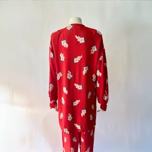 May include: A red two-piece pajama set with a repeating pattern of white floral and square designs. The long-sleeved top and pants are displayed on a mannequin. The cuffs of the top have small, decorative buttons.