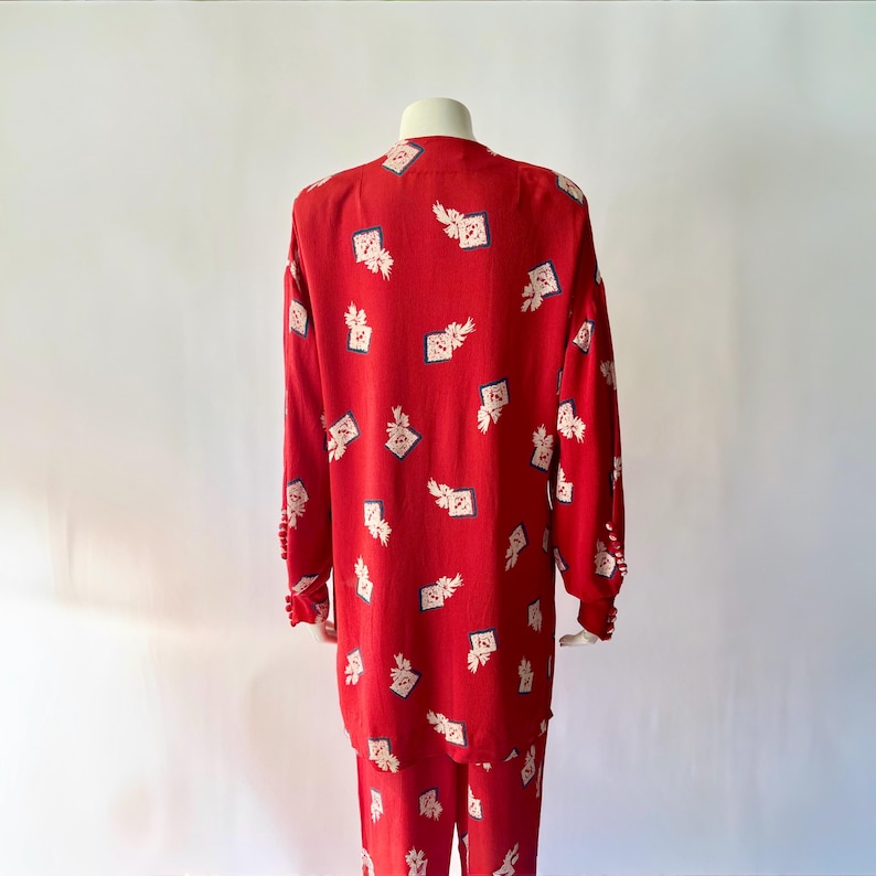 May include: A red two-piece pajama set with a repeating pattern of white floral and square designs. The long-sleeved top and pants are displayed on a mannequin. The cuffs of the top have small, decorative buttons.