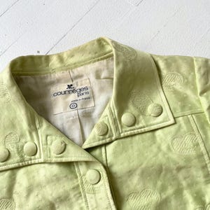 Puede incluir: Primer plano de una chaqueta verde claro con patrones en forma de coraz&oacute;n. La chaqueta tiene cuello, botones y la etiqueta "courreges paris made in france". La tela tiene una textura sutil.