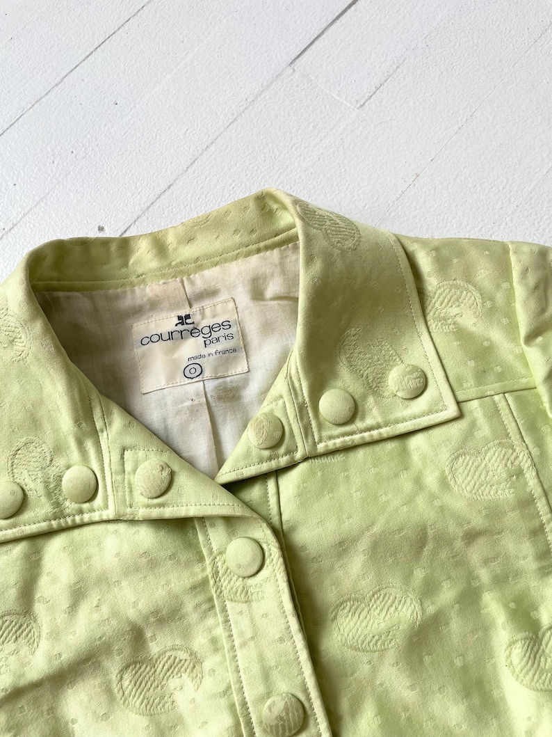 Puede incluir: Primer plano de una chaqueta verde claro con patrones en forma de coraz&oacute;n. La chaqueta tiene cuello, botones y la etiqueta "courreges paris made in france". La tela tiene una textura sutil.