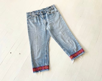 May include: Light blue denim jeans with a frayed hem and colorful embroidered trim around the bottom of the legs.