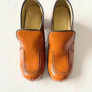 May include: A pair of brown leather slip-on shoes with black trim and stitching. The shoes have a rounded toe and a chunky heel.