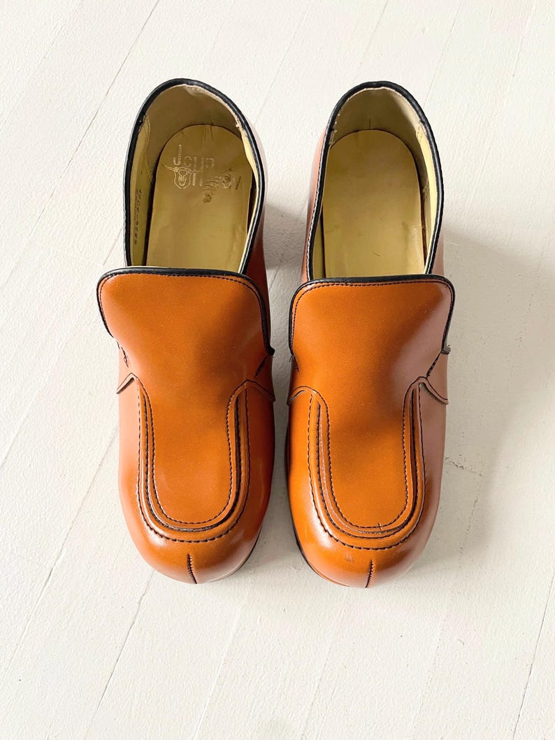 May include: A pair of brown leather slip-on shoes with black trim and stitching. The shoes have a rounded toe and a chunky heel.