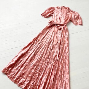 May include: A long, pink satin dress with a V-neckline and short puff sleeves. The dress has a fitted bodice and a flowing skirt with a delicate floral pattern.