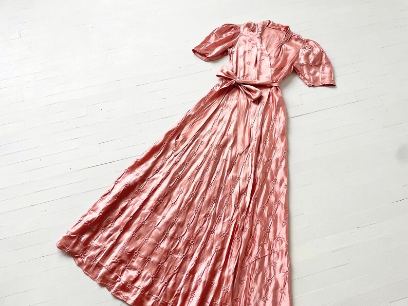 May include: A long, pink satin dress with a V-neckline and short puff sleeves. The dress has a fitted bodice and a flowing skirt with a delicate floral pattern.