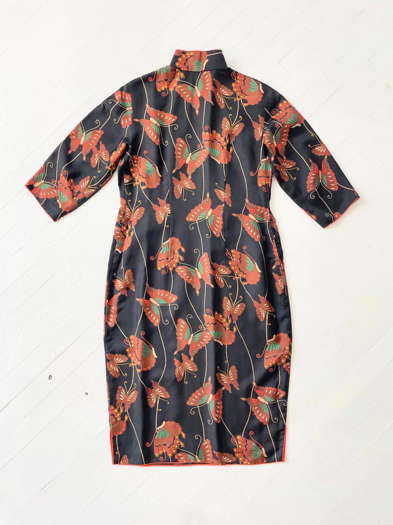 May include: A black dress with a mandarin collar and short sleeves. The dress is covered in a pattern of orange and brown butterflies on a black background. The dress has a red trim around the edges.