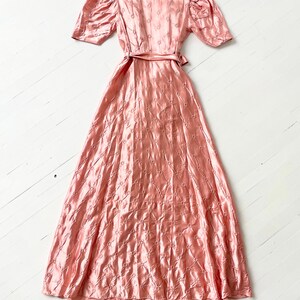 May include: A long, pink satin dress with a fitted bodice and a full skirt. The dress has a small floral pattern and a sash at the waist.