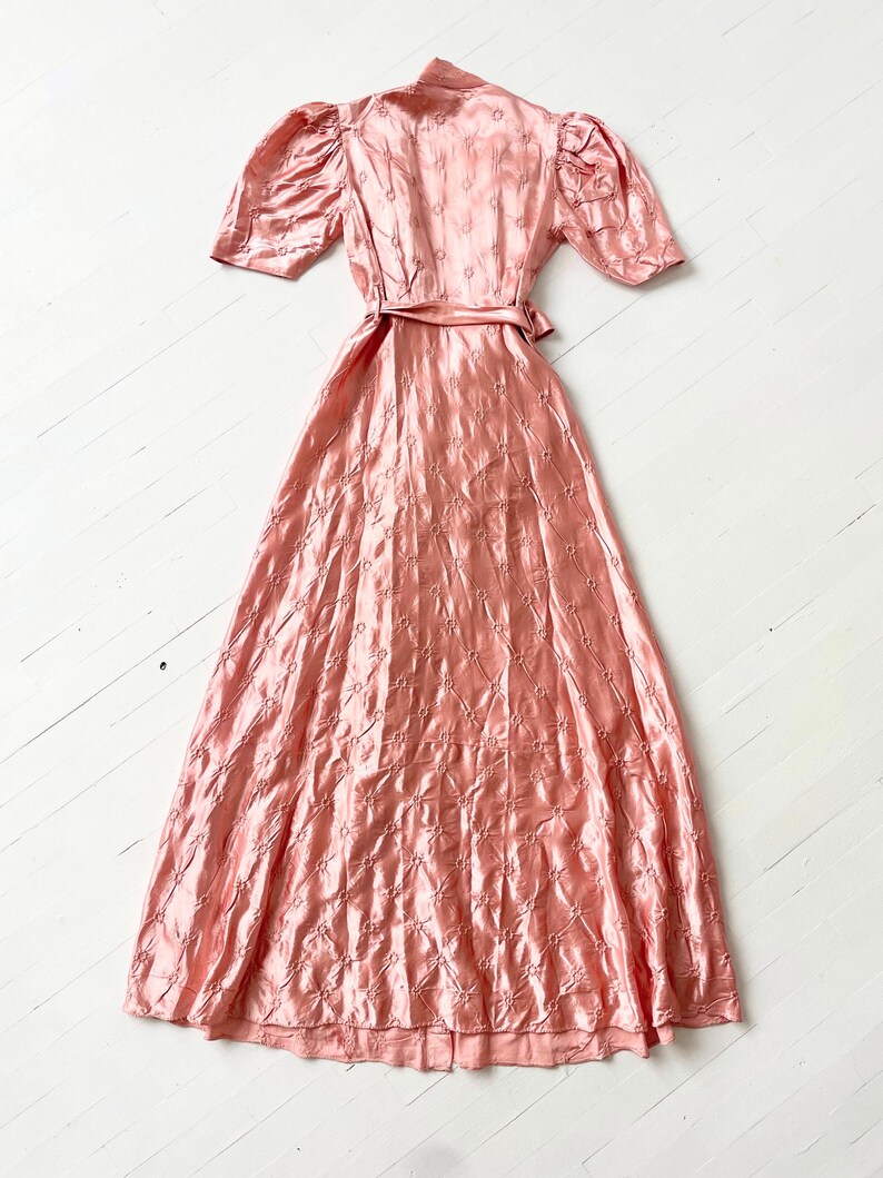May include: A long, pink satin dress with a fitted bodice and a full skirt. The dress has a small floral pattern and a sash at the waist.