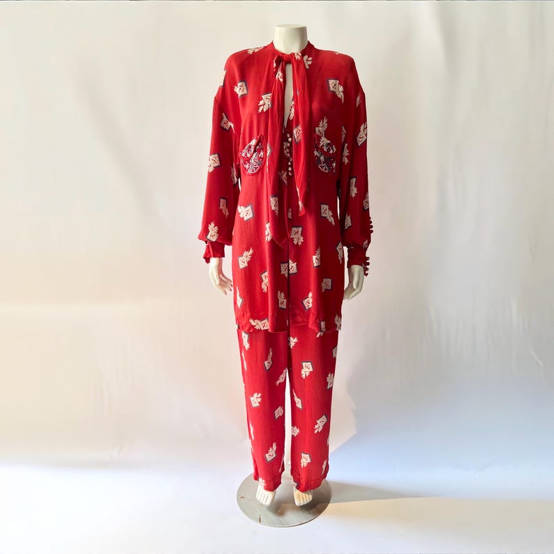 May include: A red two-piece outfit with a white pattern, displayed on a mannequin. The set includes a long-sleeved top with a tie-neck and a pair of matching pants. The top features decorative embellishments on the pockets and cuffs.