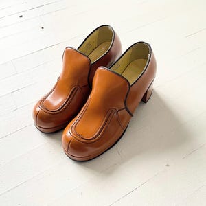 May include: A pair of brown leather slip-on shoes with a chunky heel. The shoes have a simple design with a single seam across the toe and a small flap on the top.