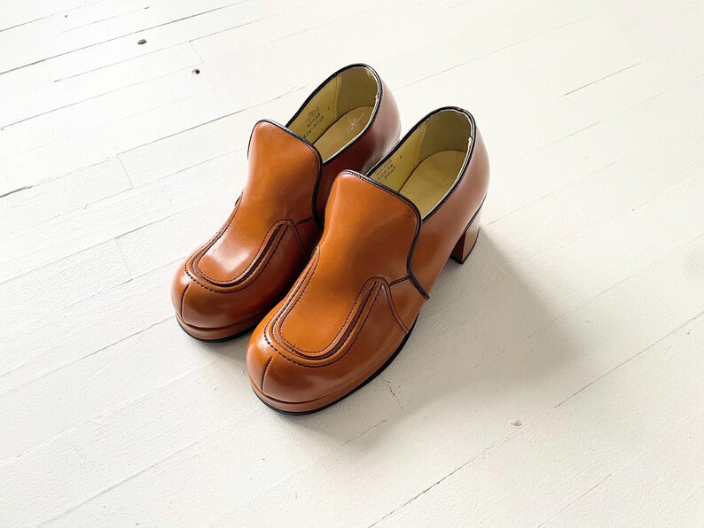 May include: A pair of brown leather slip-on shoes with a chunky heel. The shoes have a simple design with a single seam across the toe and a small flap on the top.