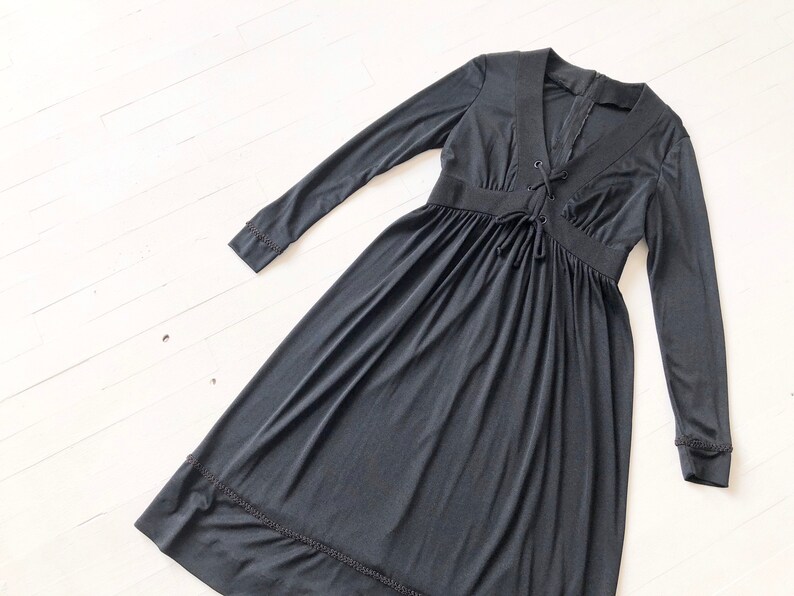 May include: A black long-sleeved dress with a V-neck and a lace-up detail at the neckline. The dress has a gathered skirt and a scalloped hem.