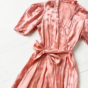 May include: A pink satin dress with a floral pattern and a self-tie sash. The dress has a V-neckline and short puff sleeves.