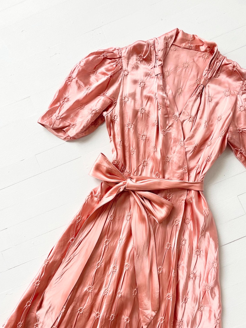 May include: A pink satin dress with a floral pattern and a self-tie sash. The dress has a V-neckline and short puff sleeves.