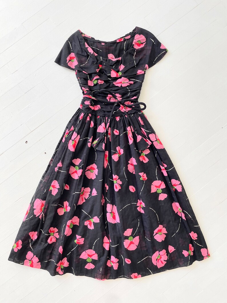 Peut inclure: Une robe vintage noire &agrave; imprim&eacute; floral rose. La robe a un corsage ajust&eacute; avec un d&eacute;collet&eacute; en c&oelig;ur et une jupe ample. La robe est cintr&eacute;e &agrave; la taille par un ruban noir.
