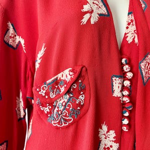 May include: Red vintage dress with white and teal floral print. The dress features a decorative pocket with intricate embroidery and a row of red and white buttons. The fabric has a textured appearance.