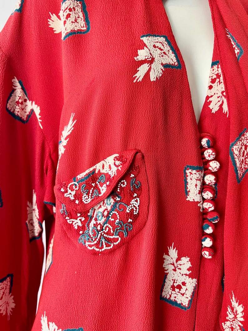 May include: Red vintage dress with white and teal floral print. The dress features a decorative pocket with intricate embroidery and a row of red and white buttons. The fabric has a textured appearance.