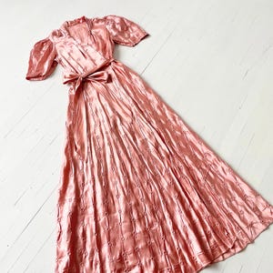 May include: A long, pink satin dress with a wrap style bodice and short puff sleeves. The dress has a delicate floral pattern and a sash tied at the waist.