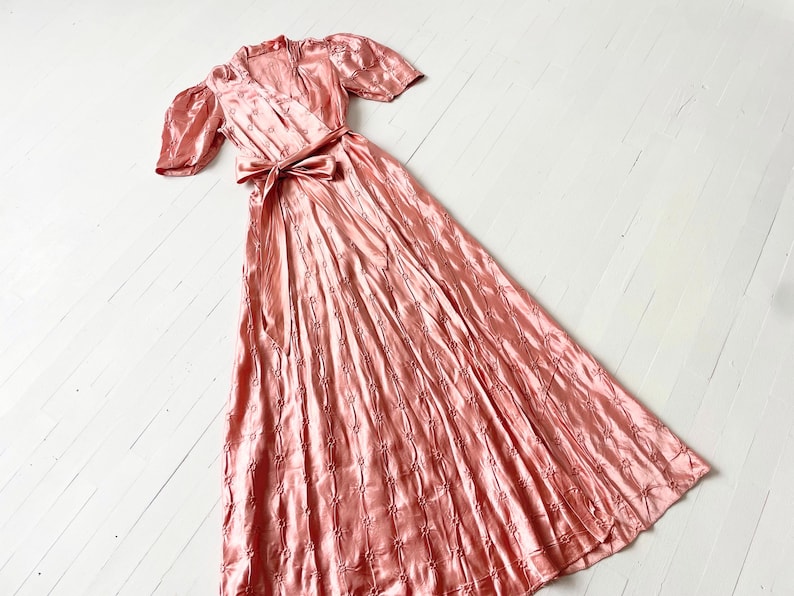 May include: A long, pink satin dress with a wrap style bodice and short puff sleeves. The dress has a delicate floral pattern and a sash tied at the waist.