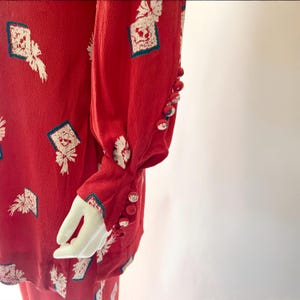 May include: Red vintage blouse with white and teal geometric print. The long-sleeved blouse features a series of small, round buttons on the cuffs. The fabric has a textured appearance.
