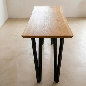 Op de afbeelding: Een houten tafel met een natuurlijke houtnerf afwerking. Het tafelblad heeft een live edge design en wordt ondersteund door vier zwarte metalen poten. De tafel is ongeveer 152 cm lang.