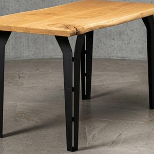 Può includere: Tavolo in legno con piano rettangolare marrone chiaro e gambe in metallo nero. Il tavolo ha una venatura naturale del legno e un bordo vivo su un lato. Le gambe sono angolate e hanno un design moderno.