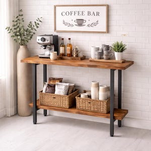 Può includere: Un mobile bar in legno per il caffè con struttura in metallo nero, dotato di macchina per il caffè, tazze e un cartello con la scritta "Coffee Bar". Il ripiano inferiore contiene cesti in vimini pieni di prodotti per il caffè.