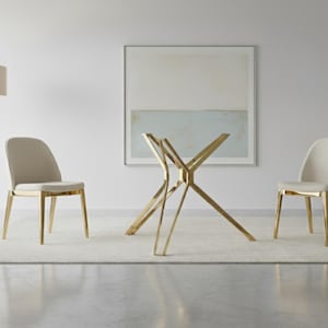 May include: A modern dining set featuring a gold-colored, starburst-style table base. Two chairs with beige upholstery and gold trim flank the table. The setting is on a light-colored rug, with a framed abstract painting in the background.