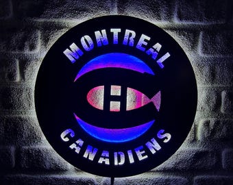 May include: A round, black sign with the words "MONTREAL CANADIENS" in white lettering. The sign features the team's logo, a stylized "H" inside a circle, with blue and pink neon lighting. The sign is mounted on a brick wall.