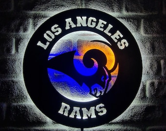 May include: A black circular sign with the words "LOS ANGELES RAMS" in white. The sign features the team's logo in blue and gold, illuminated with a white light against a brick wall background.