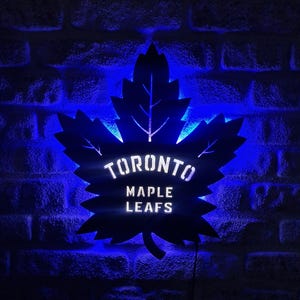 Puede incluir: Decoración de pared en forma de hoja de arce negra con el texto "TORONTO MAPLE LEAFS" en blanco. La decoración está iluminada con luces LED azules, creando un efecto brillante contra una pared de ladrillos oscuros.
