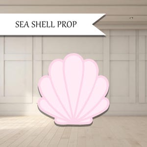 May include: A light pink seashell prop with a white banner that reads "SEA SHELL PROP". The seashell is a light pink color with a white outline and a shadow. The background is a light wood floor and white wall.