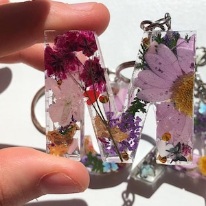 May include: A clear resin letter "M" keyring, filled with dried flowers in pink, purple, and gold. Other similar keyrings are visible in the background. The letter is held up by a hand.