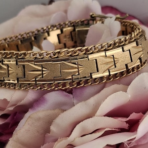 May include: A gold-toned bracelet with a geometric design and chain accents. The bracelet is resting on a bed of pink and white flowers. The bracelet's rectangular links feature a repeating arrow-like pattern.