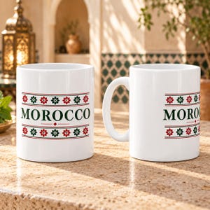 Könnte beinhalten: Zwei weiße Keramikbecher mit dem Wort MOROCCO in dunkelgrünen Großbuchstaben, zentriert zwischen zwei horizontalen Bändern aus sich wiederholenden roten und grünen Blumensternmustern und dünnen roten Linien, mit einem Henkel am rechten Becher.