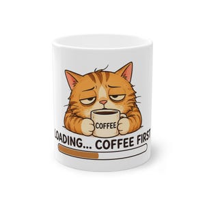 Könnte beinhalten: Ein weißer Keramikbecher mit einer Grafik einer schläfrigen, orangefarbenen getigerten Katze, die eine kleine Tasse mit der Aufschrift COFFEE hält. Darunter steht LOADING... COFFEE FIRST neben einem braun-weißen Ladebalken.