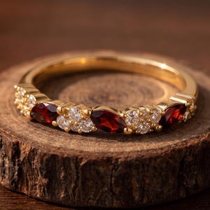 May include: A gold band ring featuring alternating marquise-cut deep red garnet stones and clusters of small round clear white stones. The ring has a polished metallic finish and is designed with a repeating pattern of gemstones along the band.