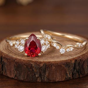 Op de afbeelding: Een gouden ringenset bestaande uit een ring met een peervormige rode edelsteen in het midden, geflankeerd door kleine ronde witte stenen, en een bijpassende gebogen gouden ring bezet met kleine ronde witte stenen langs de bovenrand.