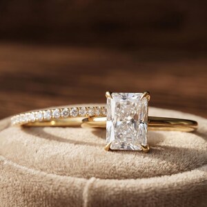 May include: A gold ring set featuring a radiant-cut centre stone held by four prongs on a plain gold band, accompanied by a matching thin gold band encrusted with a row of small, round, clear stones along the top half of the ring's circumference.